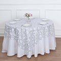 Round white tablecloth with silver floral design on a dining table.