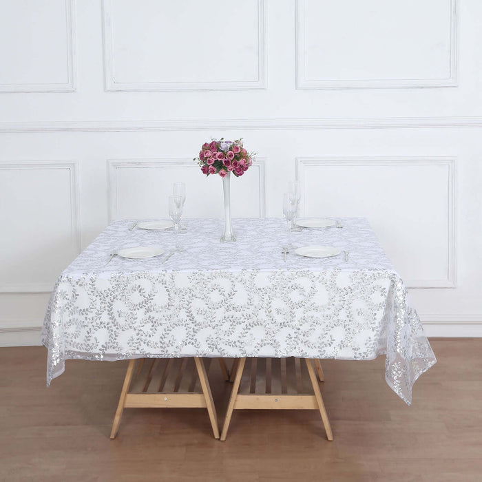 White lace tablecloth on a wooden table set for dining with flowers and glassware.
